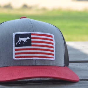 May include: A gray and red baseball cap with a black mesh back. The cap features a white embroidered American flag with a white silhouette of a dog running in the top left corner.
