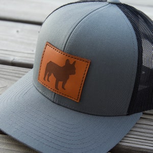 Puede incluir: Gorra trucker gris con parte trasera de malla negra y parche de cuero marr&oacute;n con la silueta de un bulldog franc&eacute;s. La gorra tiene una visera curva y cierre snapback.