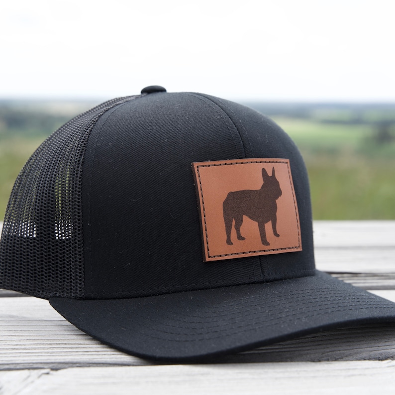Puede incluir: Gorra de camionero negra con un parche de cuero marr&oacute;n con la silueta de un bulldog franc&eacute;s. La gorra tiene una parte trasera de malla y una visera curva. El parche rectangular est&aacute; cosido en la parte delantera.