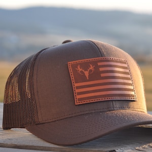 K&ouml;nnte beinhalten: Eine braune und schwarze Trucker-Kappe mit einem braunen Leder-Patch, der eine amerikanische Flagge mit einer Hirschkopf-Silhouette in der Mitte zeigt.