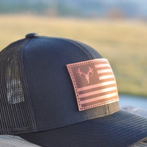 K&ouml;nnte beinhalten: Eine schwarze und braune Trucker-Kappe mit einem Leder-Patch, der eine stilisierte amerikanische Flagge mit einem Hirschsch&auml;del in der Mitte zeigt.