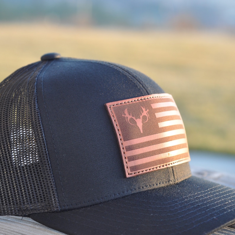 K&ouml;nnte beinhalten: Eine schwarze und braune Trucker-Kappe mit einem Leder-Patch, der eine stilisierte amerikanische Flagge mit einem Hirschsch&auml;del in der Mitte zeigt.