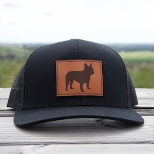 Puede incluir: Gorra de camionero negra con un parche de cuero marr&oacute;n con la silueta de un bulldog franc&eacute;s. La gorra tiene una parte trasera de malla y una visera curva. La gorra est&aacute; sobre una superficie de madera.