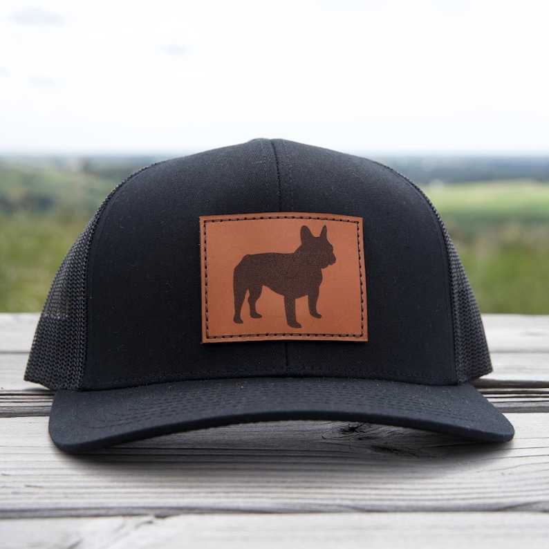 Puede incluir: Gorra de camionero negra con un parche de cuero marr&oacute;n con la silueta de un bulldog franc&eacute;s. La gorra tiene una parte trasera de malla y una visera curva. La gorra est&aacute; sobre una superficie de madera.