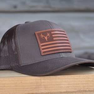 K&ouml;nnte beinhalten: Eine braune und schwarze Mesh-Trucker-Kappe mit einem braunen Leder-Patch, der eine amerikanische Flagge mit einem Hirschkopf in der Mitte zeigt.