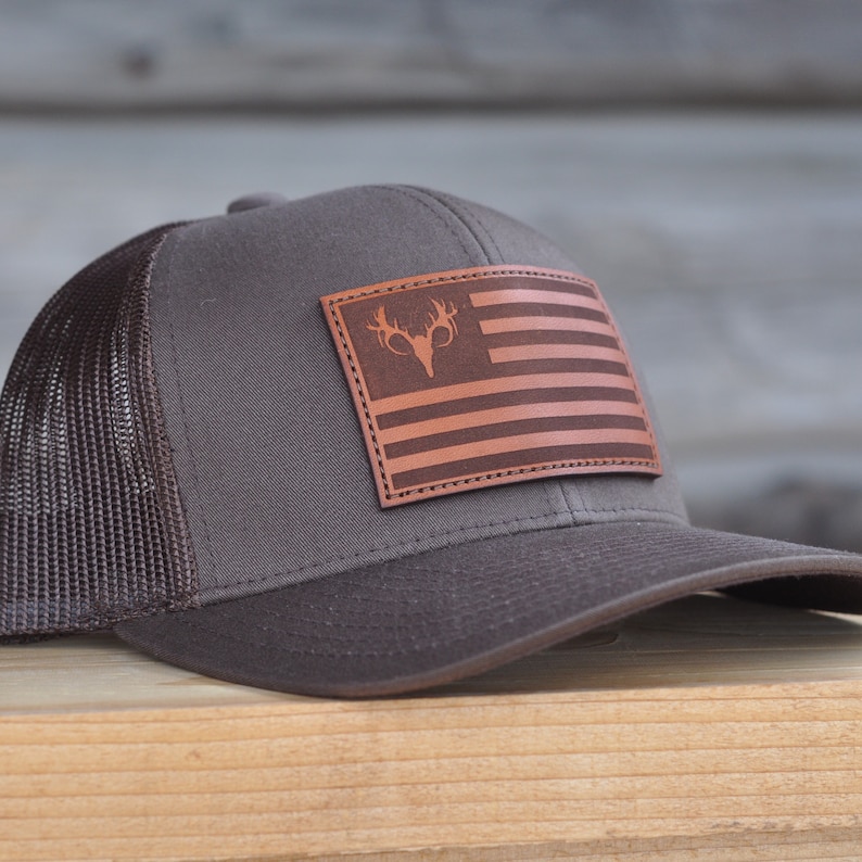 K&ouml;nnte beinhalten: Eine braune und schwarze Mesh-Trucker-Kappe mit einem braunen Leder-Patch, der eine amerikanische Flagge mit einem Hirschkopf in der Mitte zeigt.