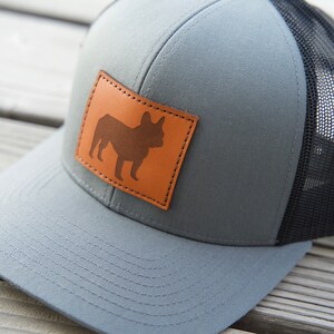Puede incluir: Gorra de camionero gris con parte trasera de malla negra y parche de cuero marr&oacute;n con silueta de bulldog franc&eacute;s. La gorra tiene visera curva y cierre snapback, adecuada para uso casual.