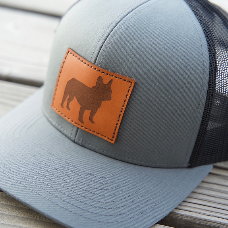 Puede incluir: Gorra de camionero gris con parte trasera de malla negra y parche de cuero marr&oacute;n con silueta de bulldog franc&eacute;s. La gorra tiene visera curva y cierre snapback, adecuada para uso casual.