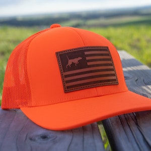 May include: Bright orange trucker hat with a brown leather patch featuring an American flag design with a dog silhouette. The hat has a mesh back and a curved brim. A stylish accessory for outdoor enthusiasts.