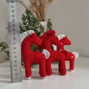 May include: Three red crocheted horse figurines with silver manes and tails, arranged on a white surface. The tallest horse measures approximately 13 cm in height. Decorative dried flowers and greenery are in the background.