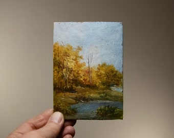 Op de afbeelding: Een klein olieverfschilderij dat een landschapsscene met een beek en bomen in herfstkleuren afbeeldt. De bomen zijn een mix van geel, oranje en bruin, en de beek is lichtblauw. De lucht is lichtblauw met wat witte wolken.