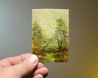 Op de afbeelding: Een klein schilderij op canvas dat een landschapsscene met bomen en een groen veld voorstelt. Het schilderij heeft een vergeelde achtergrond en een rustieke, vintage esthetiek.