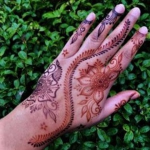 May include: A hand with intricate henna designs in shades of brown and black. The design features a large flower with a circular center and swirling patterns.