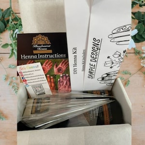 Könnte beinhalten: Eine weiße Box mit einem DIY-Henna-Set. Die Box enthält Hennapaste, Konen und Anweisungen. Die Box enthält auch eine Karte mit Beispielen für Henna-Designs.