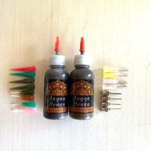 May include: Two bottles of black henna paste labeled "Jagua Henna" with red caps and orange mandala designs on the labels. The bottles are surrounded by a variety of colored plastic applicators and metal needles.