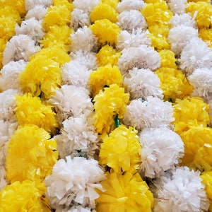 May include: A dense arrangement of artificial marigold flowers in alternating bright yellow and white colors. The flowers are constructed from thin, layered plastic strips, creating a full, rounded, and textured appearance across the entire display.