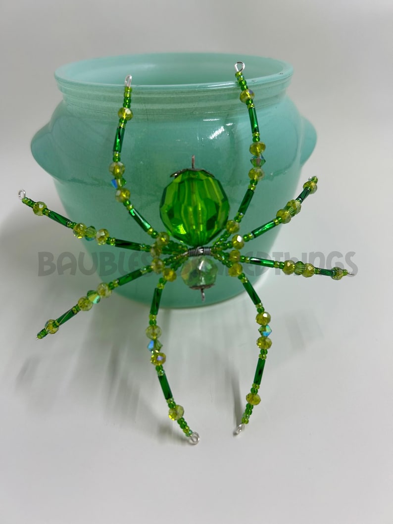 May include: A green beaded spider ornament with a crystal body and iridescent green beads. The spider is sitting on top of a light blue ceramic jar.