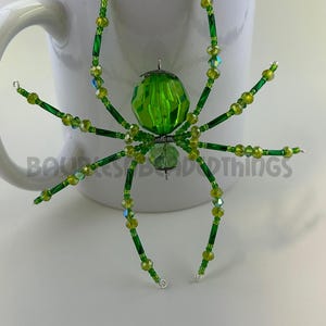 May include: A green beaded spider ornament with a large green crystal body. The spider has eight legs made of green beads and silver wire.