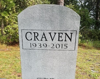 May include: A grey tombstone with the name "CRAVEN" and the dates "1939-2015" inscribed on it. The inscription "MAY THE NIGHTMARES NEVER END" is also visible on the tombstone.