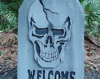 May include: A grey, cracked, tombstone-shaped Halloween decoration with a black skull and the word "WELCOME" printed in black.