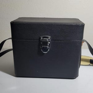 May include: Black rectangular camera case with a textured surface, metal clasp, and a shoulder strap. The case is boxy and appears to be made of leather or a leather-like material. The strap is black with a yellow and black accent.