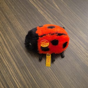 May include: A plush ladybug toy with a red body and black spots. The toy has a black head and small legs. A yellow tag with the word "Cappy" and other text is attached to the toy.