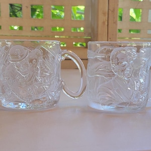 May include: Two clear glass mugs with detailed embossed designs. One mug features a figure in a hat, the other a face. Both have curved handles. The mugs are set against a blurred background.