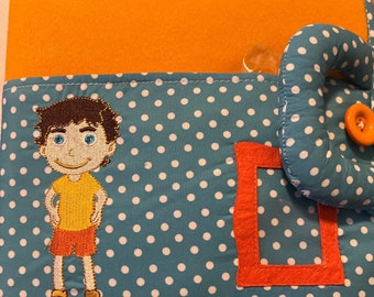 May include: A blue fabric book with white polka dots. The book features a smiling boy with brown hair, a yellow shirt, and red shorts. There is a red frame and a blue and white polka dot elephant with a red button.