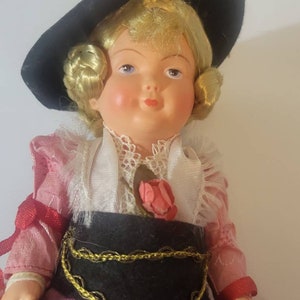 May include: A vintage porcelain doll wearing a pink and red dress with a black belt and a black hat. The doll has blonde hair and blue eyes.