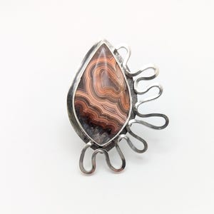 May include: A silver-toned ring featuring a teardrop-shaped stone with brown and cream-colored banded patterns. The stone is set in a silver bezel with a decorative, openwork design extending from the side.