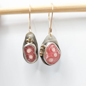 May include: A pair of handcrafted earrings featuring teardrop-shaped, silver-toned metal settings. Each earring showcases a vibrant pink stone with concentric circles, accented with gold-colored details and gold-toned ear wires.