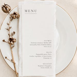 May include: A white dinner plate topped with a folded white napkin and a vertical menu card. The menu lists an entrée of heirloom tomatoes, buffalo milk mozzarella, basil, shallots, and olive oil, a main of pappardelle, and a raspberry frangipane dessert.