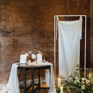 May include: A dark wooden round table draped with a white fabric runner, featuring small floral arrangements and glass candle holders. Beside it stands a white metal frame holding a long white fabric seating chart with the text Please be seated.