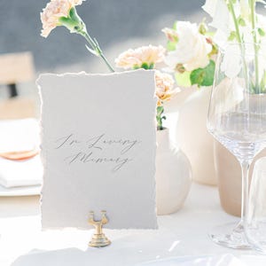 May include: A table setting with a card that reads "In Loving Memory" in elegant script. The card is held by a gold-colored metal holder. Soft peach-colored flowers and white vases are in the background, with a wine glass on the right.