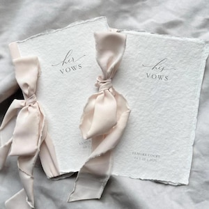 May include: Two white vow books with deckled edges, tied with blush silk ribbons. The text "her vows" and "his vows" are printed in elegant script. The date and location are printed at the bottom.