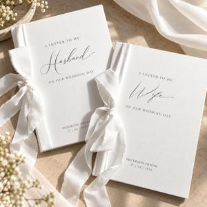 Può includere: Due libri con copertina rigida bianca e nastri bianchi. Un libro riporta la scritta A LETTER TO MY Husband ON OUR WEDDING DAY, l'altro A LETTER TO MY Wife ON OUR WEDDING DAY. Entrambi presentano PETERSON HOUSE 27 | 04 | 2024 in testo nero.