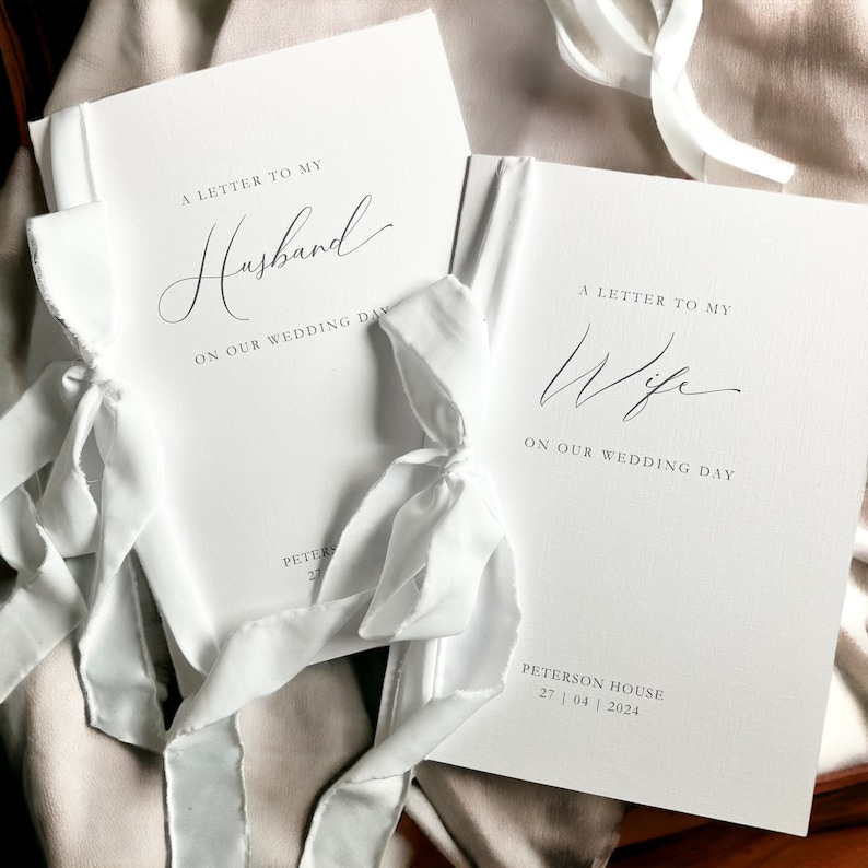 May include: Two white books with white ribbon ties. The covers have the text "A Letter to My Husband" and "A Letter to My Wife" with the date "27 | 04 | 2024" on the bottom right corner.