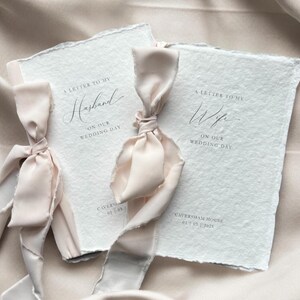 Pu&ograve; includere: Due libretti in carta bianca testurizzata con bordi irregolari, ciascuno legato con un nastro di seta rosa pallido. Il libretto a sinistra riporta A LETTER TO MY Husband ON OUR WEDDING DAY, quello a destra A LETTER TO MY Wife ON OUR WEDDING DAY, entrambi con Caversham House 01 03 2025.