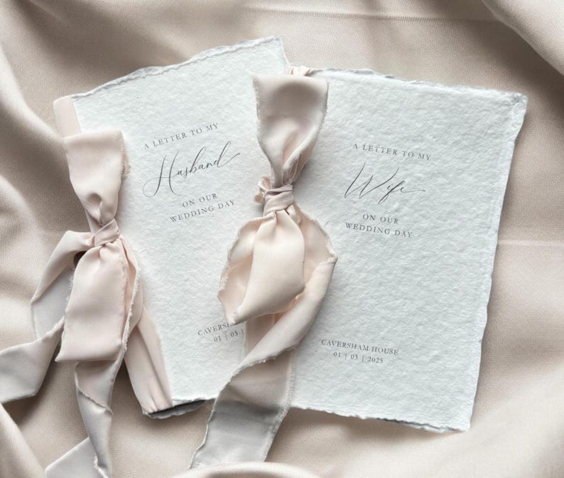 Pu&ograve; includere: Due libretti in carta bianca testurizzata con bordi irregolari, ciascuno legato con un nastro di seta rosa pallido. Il libretto a sinistra riporta A LETTER TO MY Husband ON OUR WEDDING DAY, quello a destra A LETTER TO MY Wife ON OUR WEDDING DAY, entrambi con Caversham House 01 03 2025.
