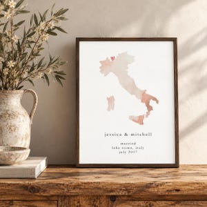 Könnte beinhalten: Ein gerahmter Kunstdruck mit einer Aquarellkarte von Italien in Pfirsich- und Beigetönen. Ein kleines rotes Herz markiert einen Ort auf der Karte. Unter der Karte steht der Text: jessica & mitchell, married, lake como, italy, july 2017.