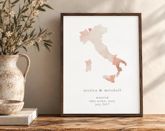 Puede incluir: Una lámina de arte enmarcada con un mapa en acuarela de Italia en tonos melocotón y bronceado. Un pequeño corazón rojo marca una ubicación en el mapa. Debajo del mapa, el texto dice: jessica & mitchell, married, lake como, italy, july 2017.