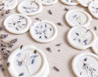 Puede incluir: Una colección de sellos de cera blancos circulares con un diseño de rama en relieve y capullos de lavanda secos incrustados. Los sellos están dispuestos en un patrón disperso junto a ramas sueltas de flores de lavanda secas.