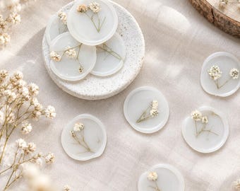 Puede incluir: Una colección de sellos de cera circulares con un acabado blanquecino translúcido, cada uno incrustado con delicadas flores secas de paniculata blanca. Varios sellos están apilados en un plato de cerámica moteado.