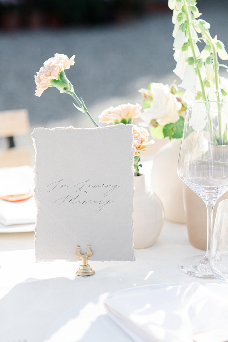 May include: A table setting with a card that reads "In Loving Memory" in elegant script. The card is held by a gold-colored metal holder. Soft peach-colored flowers and white vases are in the background, with a wine glass on the right.