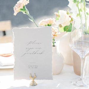 May include: A wedding guestbook sign with the text "Please sign our guestbook" in elegant script. The sign is held by a gold-colored metal holder and is surrounded by flowers and glassware, creating a romantic ambiance.