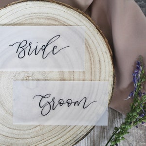 May include: Two white cards with black calligraphy on a wooden surface. The top card says "Bride" and the bottom card says "Groom".