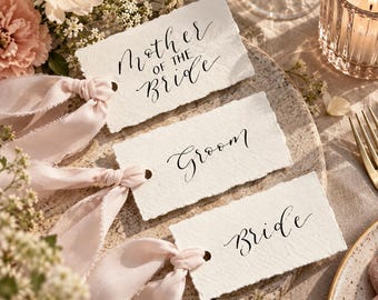 Puede incluir: Tres etiquetas rectangulares de papel blanco roto con bordes irregulares, cada una con caligrafía negra que dice Mother of the Bride, Groom y Bride. Cada etiqueta tiene un agujero perforado a la izquierda con una cinta de seda rosa pálido.