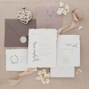 May include: A collection of wedding stationery, including invitation cards, RSVP cards, and details cards. The design features handmade paper, botanical illustrations, and elegant calligraphy. A pearl necklace, ribbon, and wax seals add to the aesthetic.