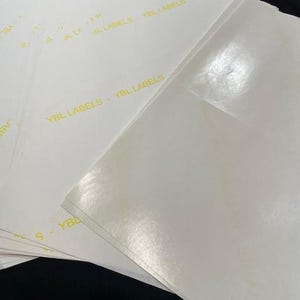 Pode incluir: Uma pilha de etiquetas retangulares brancas e brilhantes com o texto "YBL LABELS" impresso em amarelo.