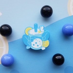 Puede incluir: Un pin de esmalte azul y blanco de un té de boba con una rodaja de limón y una perla de boba en forma de luna. El pin está rodeado de seis pequeñas canicas redondas y coloridas.
